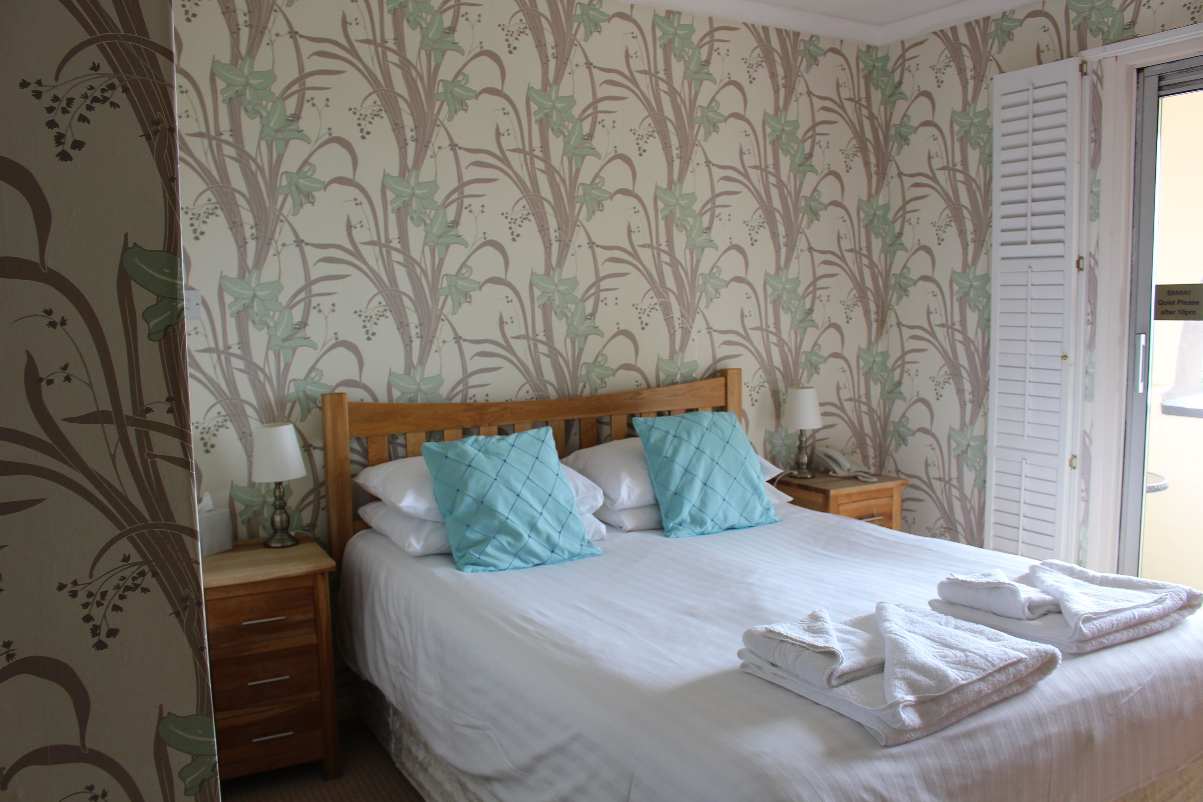 Una cama bien hecha con sábanas blancas y dos almohadas color turquesa, una mesita de noche de madera con una lámpara, y una pared con papel tapiz floral.