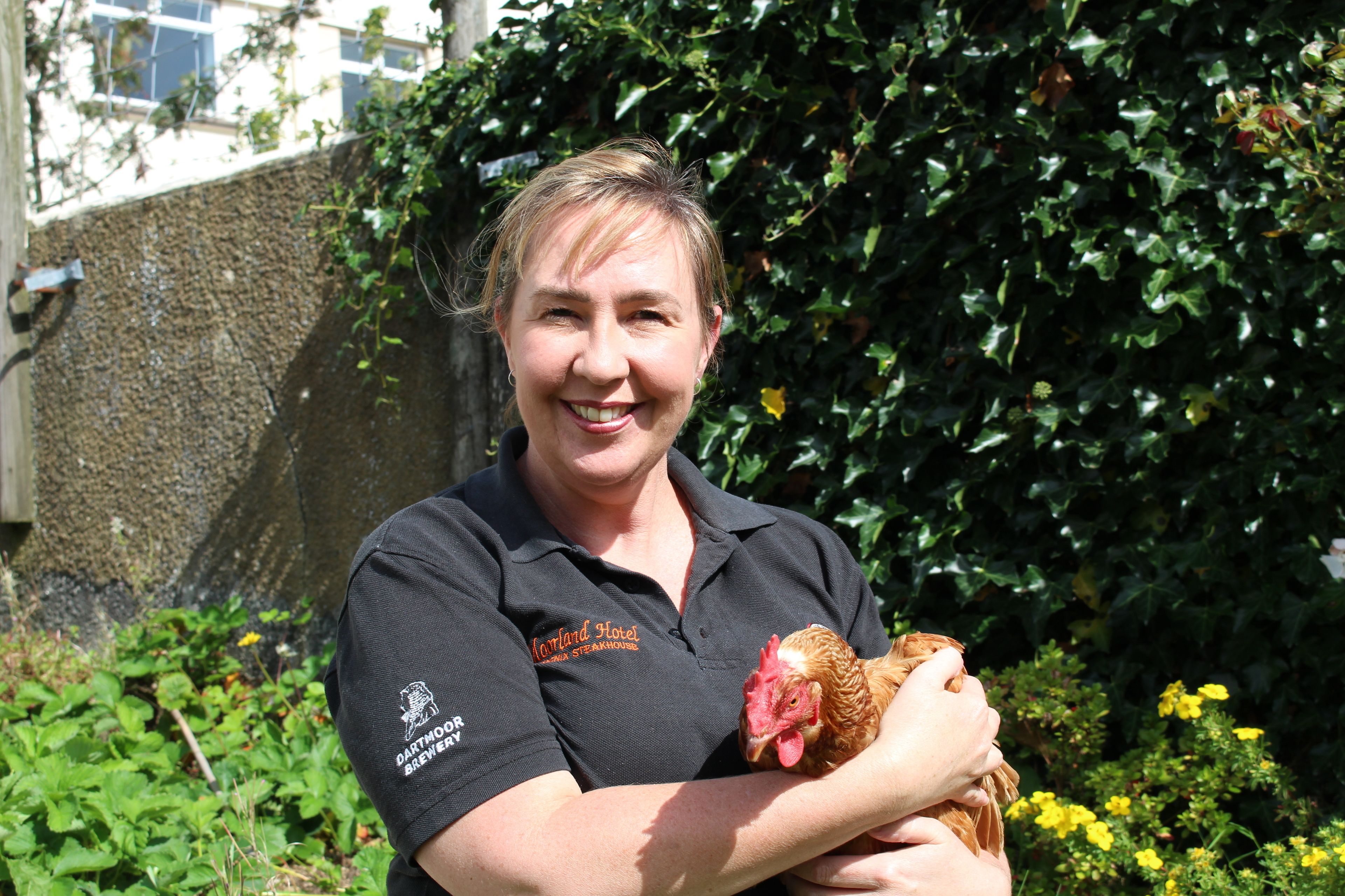 A person holding a chicken, wearing a shirt with "Dartmoor Brewery" text, standing in front of a stone wall and greenery.