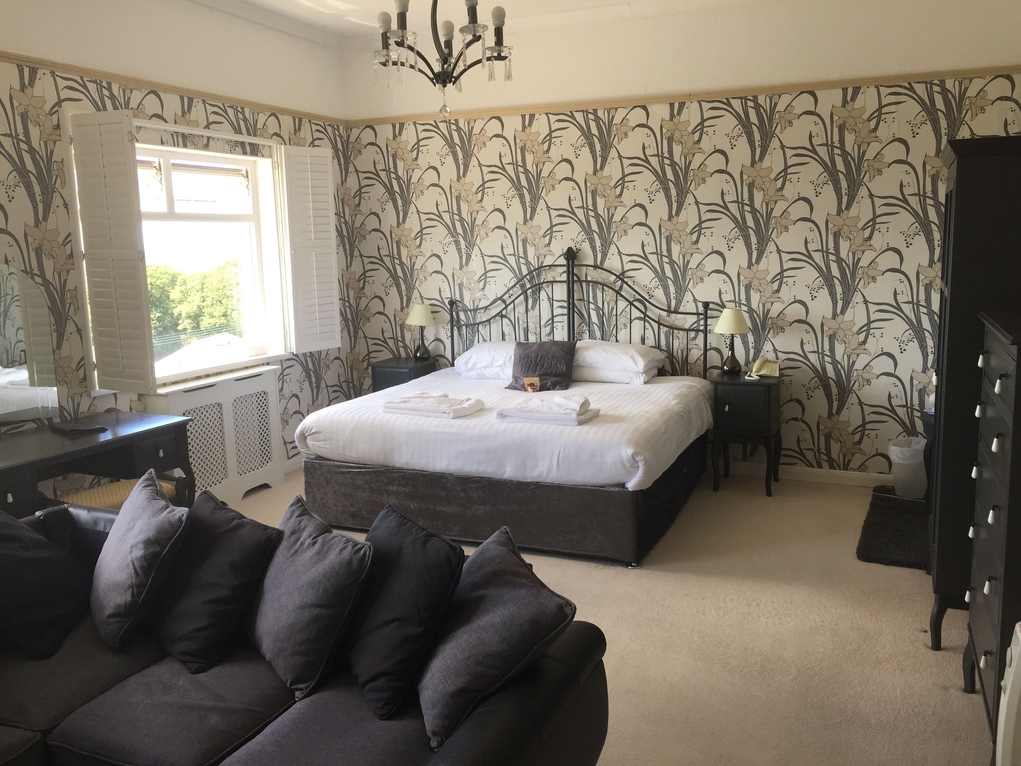 A bedroom with a large bed, a dark sofa, a chandelier, and a window with shutters.