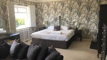 A bedroom with a large bed, a dark sofa, a chandelier, and a window with shutters.
