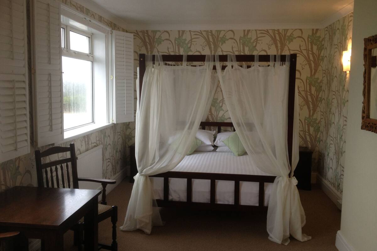A bedroom with a four-poster bed, a wooden chair, a table, and a window with shutters.