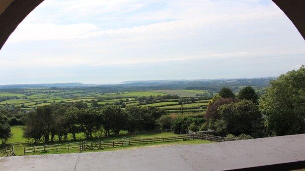A view through an arched window of a rural landscape with rolling green fields and distant hills.