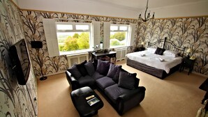 A bedroom with a large bed, a sofa, and a desk. There are two windows with shutters, and a TV is mounted on the wall.
