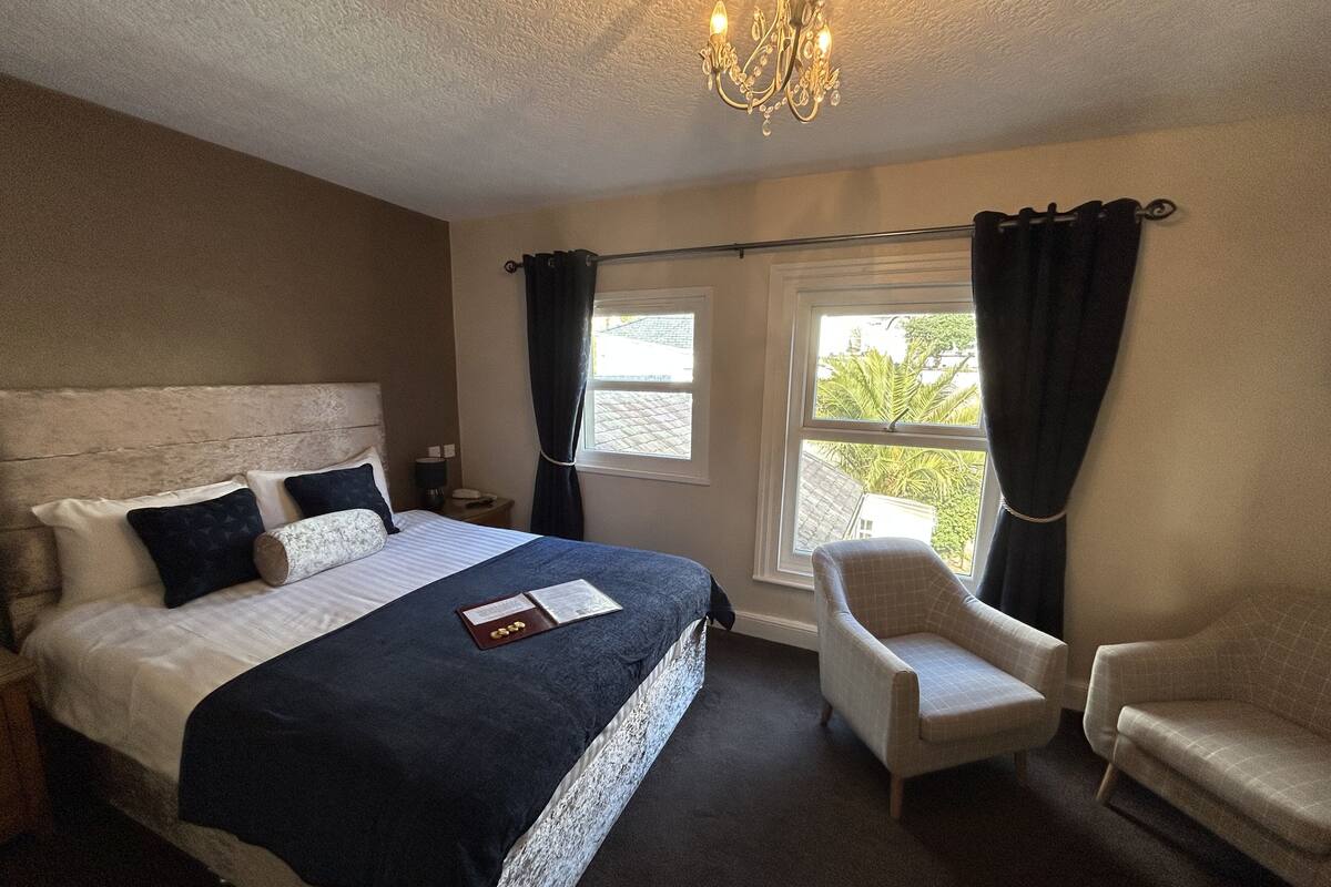 A bedroom with a large bed, two chairs, a nightstand, a chandelier, and a window with curtains.