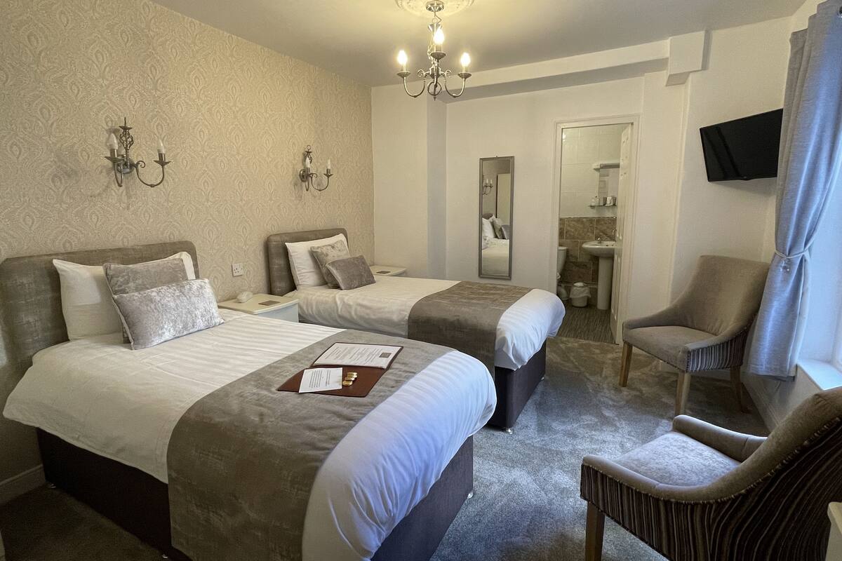 A hotel room with two beds, a sitting area with a chair, a television mounted on the wall, and a bathroom visible through an open door.