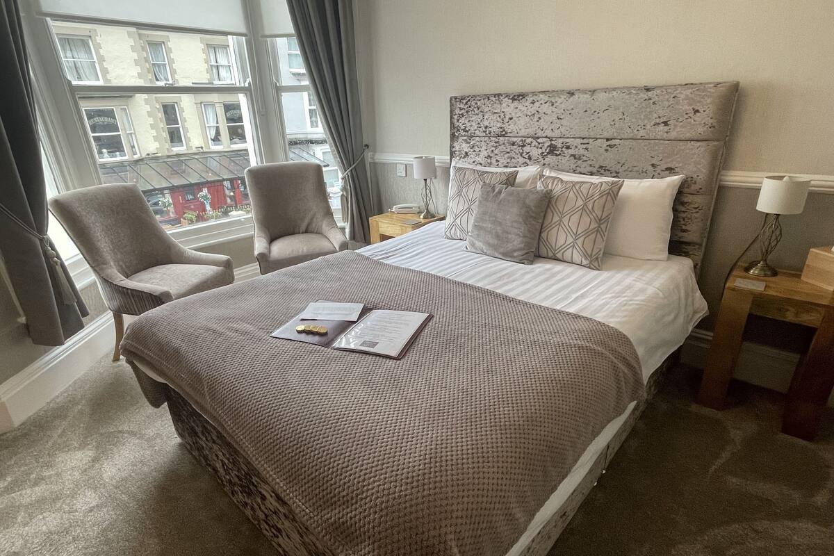 A hotel room with a large bed, two armchairs, a view of buildings outside the window, and a bedside table with a lamp.