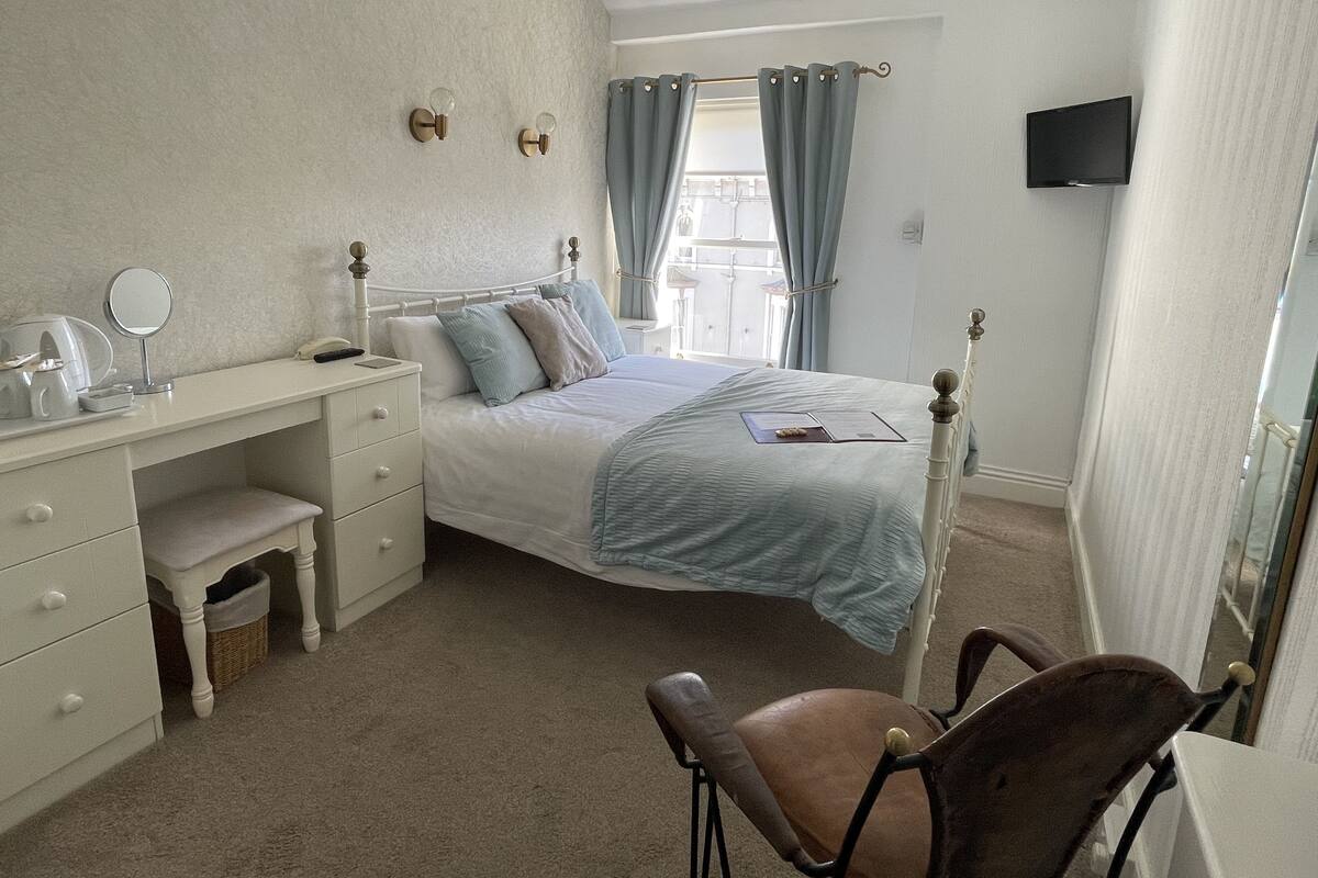 A bedroom with a bed, a desk with a mirror, a chair, and a TV on the wall.