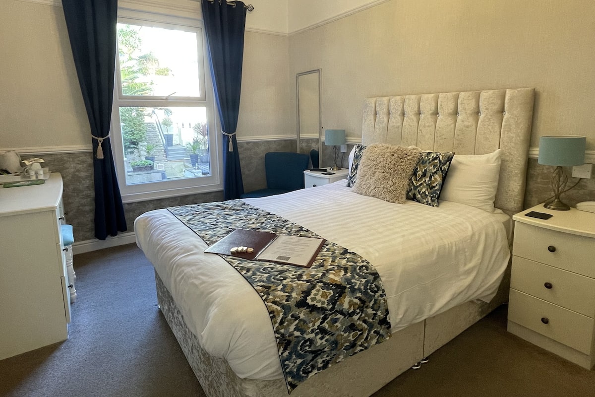 A neatly arranged bedroom with a large bed, a bedside table, a chair, and a window with curtains.