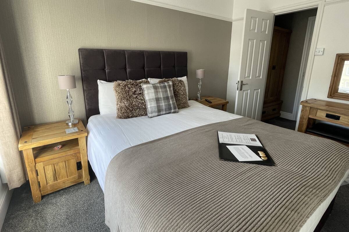 A neatly made bed with a grey bedspread, a wooden nightstand with a lamp, and a bedside table with a remote control.