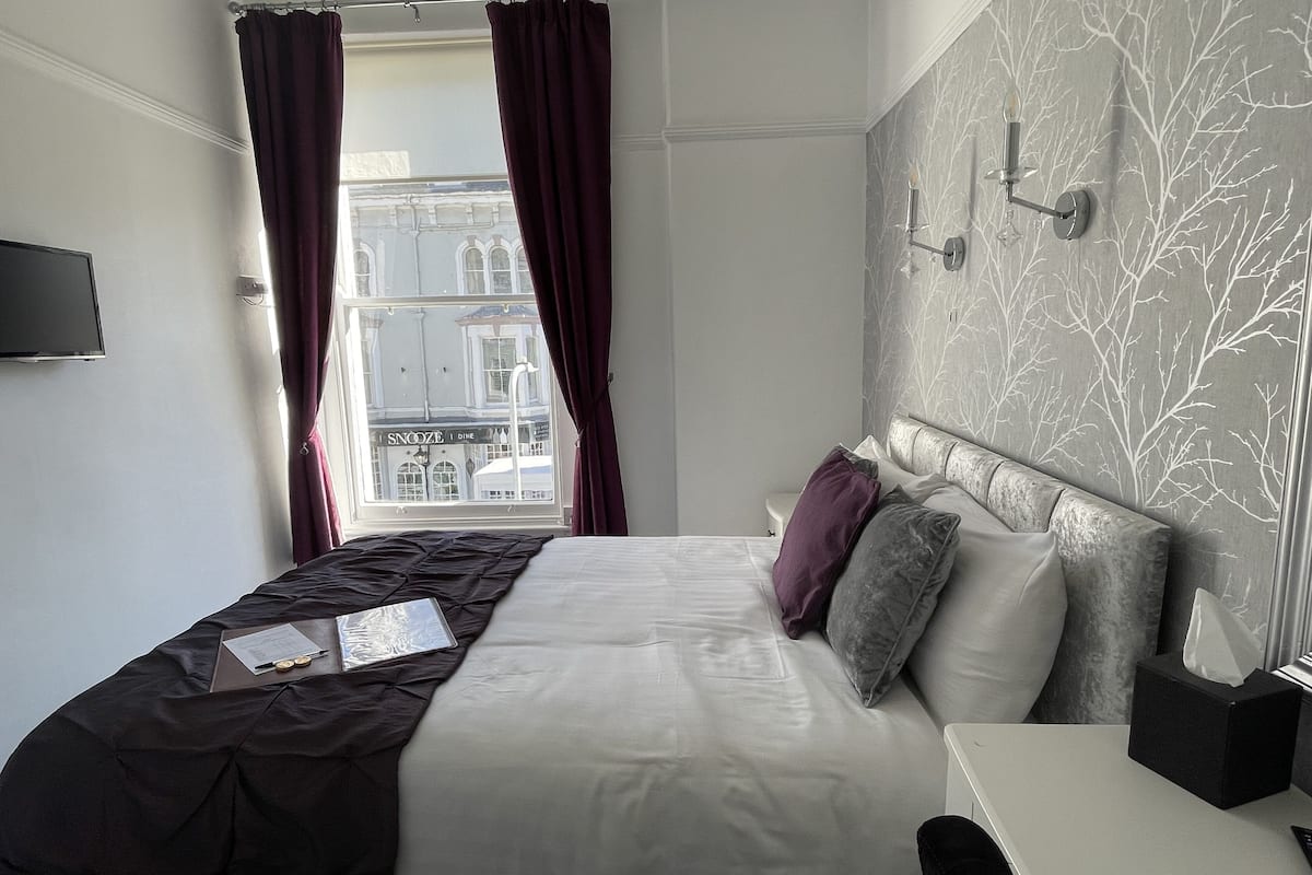 A hotel room with a bed, a bedside table, a window with curtains, and a wall-mounted light fixture.