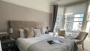 A hotel room with a large bed, a nightstand, a chair, and a view of the city through the window. - Lynton House (Llandudno)