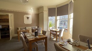 A dining area with a wooden table set for four, a chandelier, and a view of the city through large windows. - Lynton House (Llandudno)