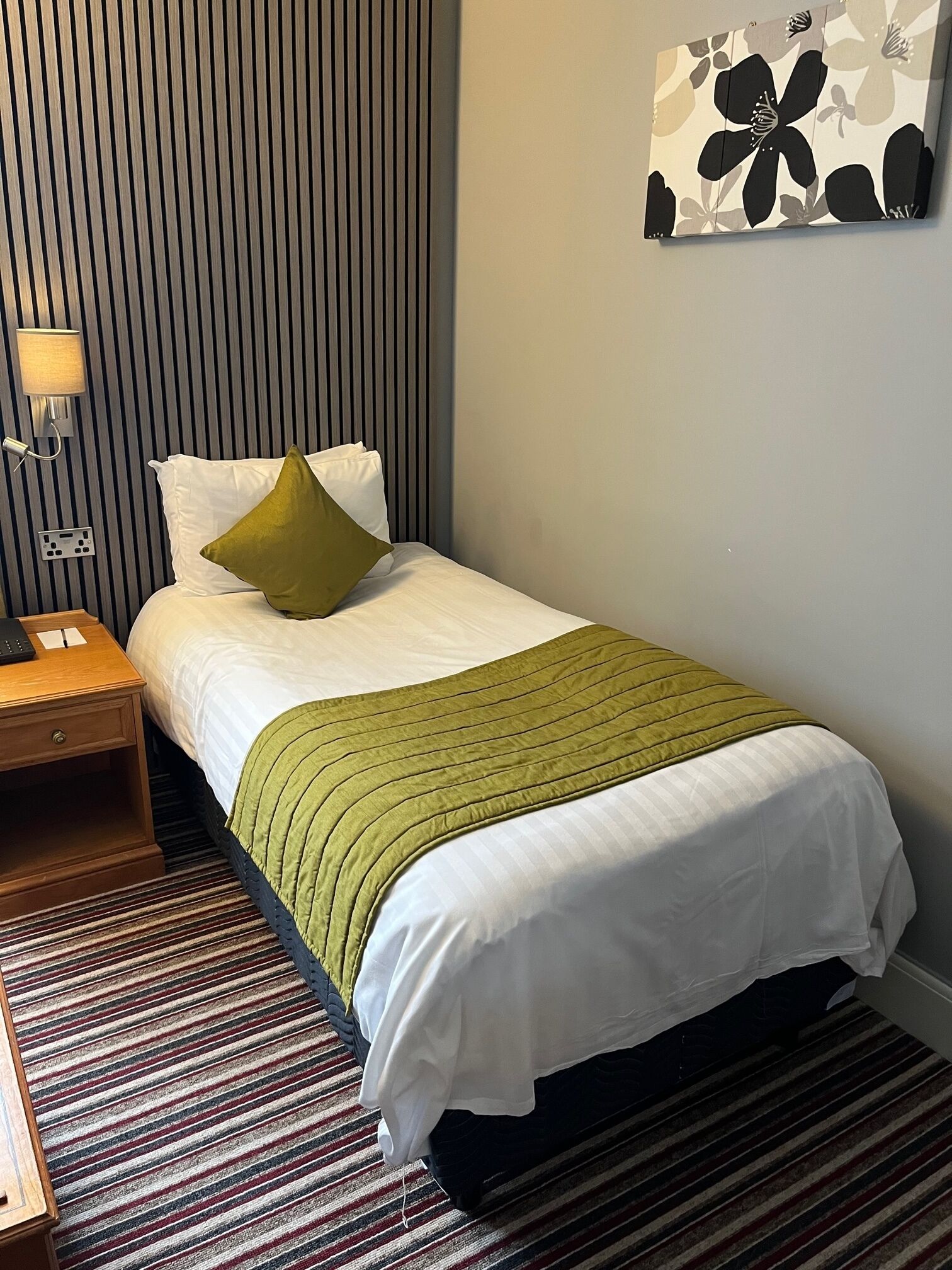 A neatly made bed with white linens and a green blanket, a bedside table with a lamp, and a striped carpet.