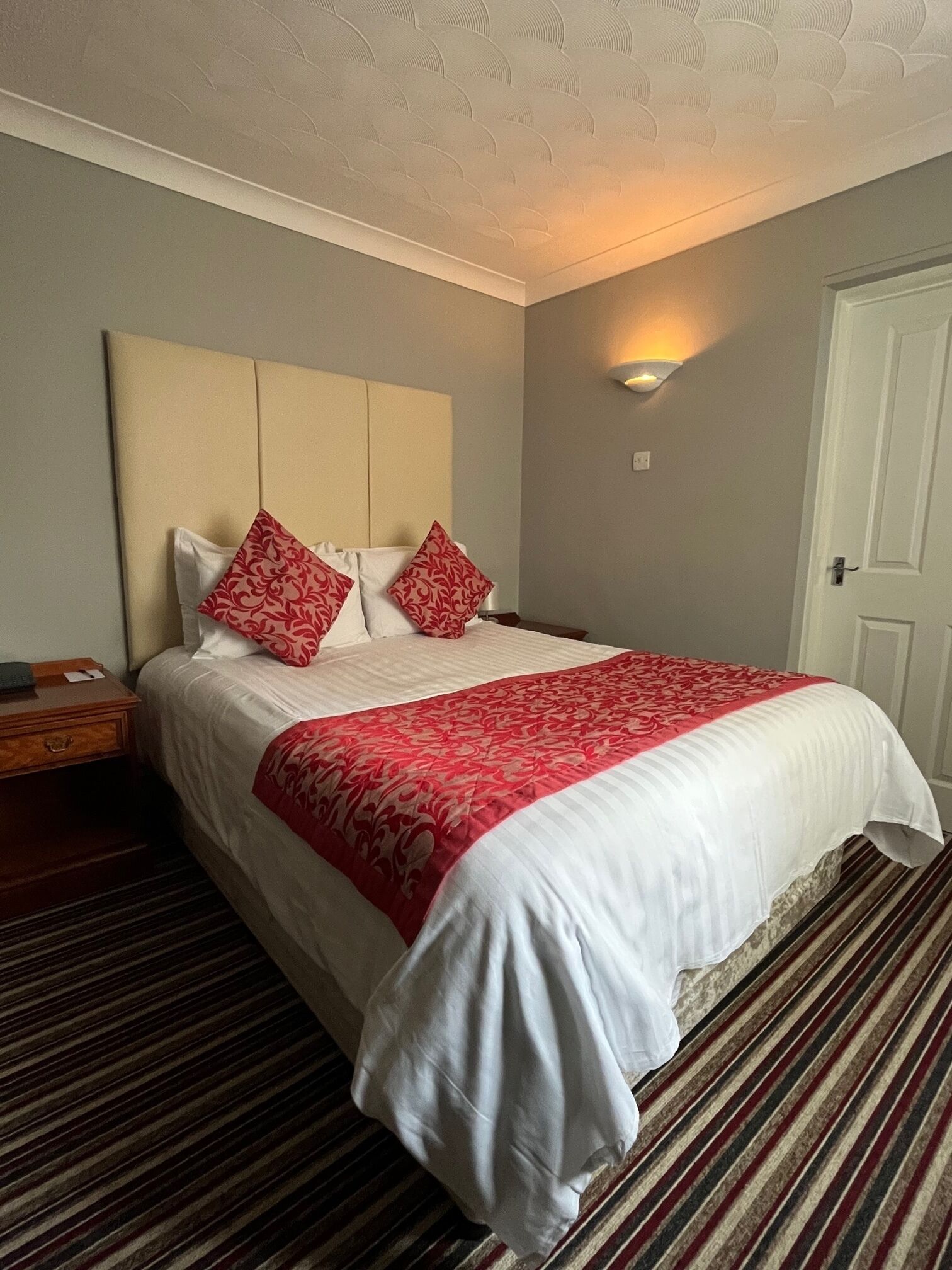 A neatly made bed with white and red bedding, a wooden nightstand, and a striped carpet.