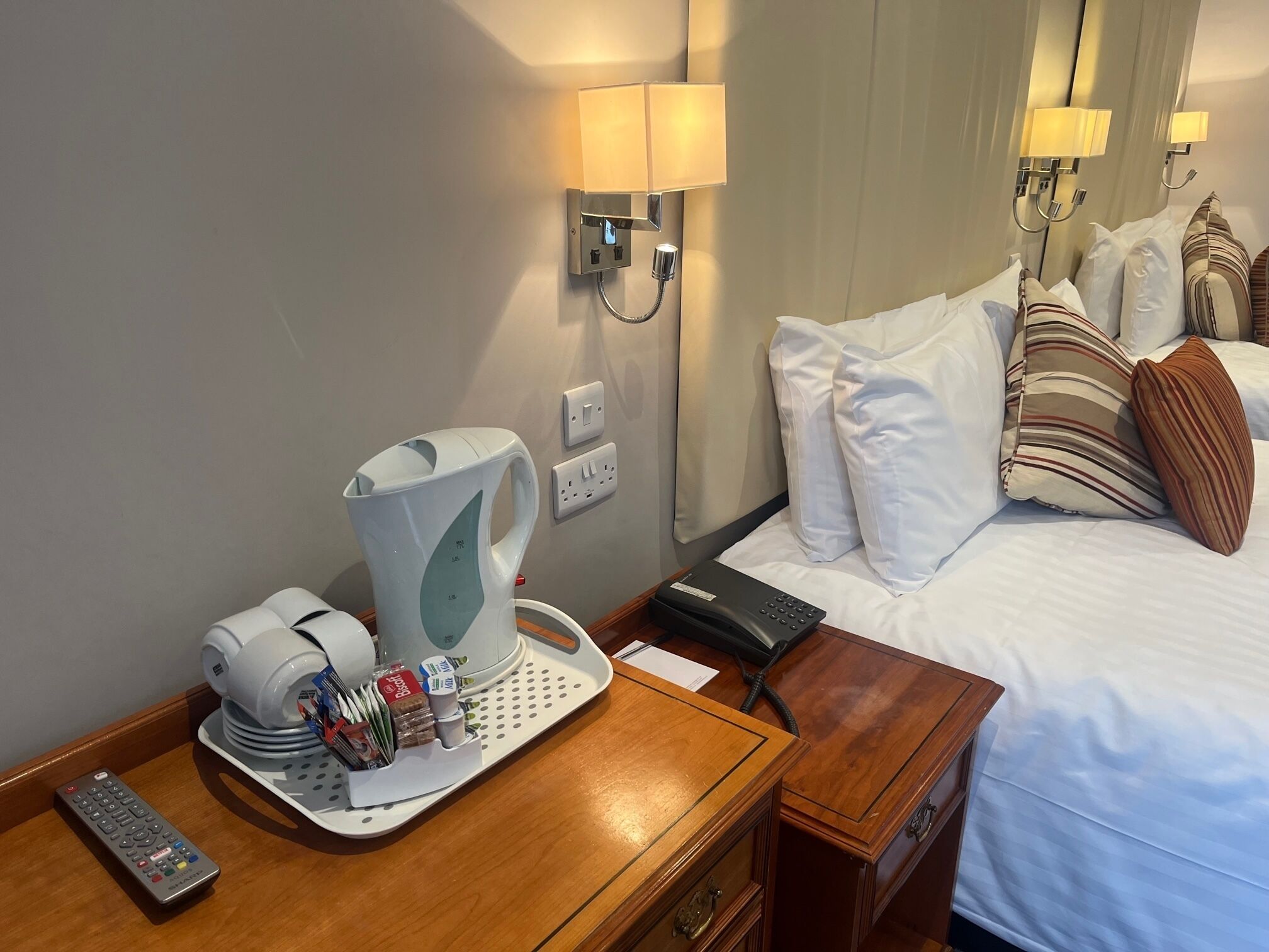 A hotel room with a bed, bedside lamps, a coffee maker, a remote control, and a tray with toiletries.
