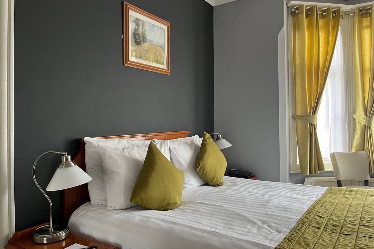 A hotel room with a bed, two pillows, a nightstand with a lamp, a framed picture on the wall, and a window with curtains.