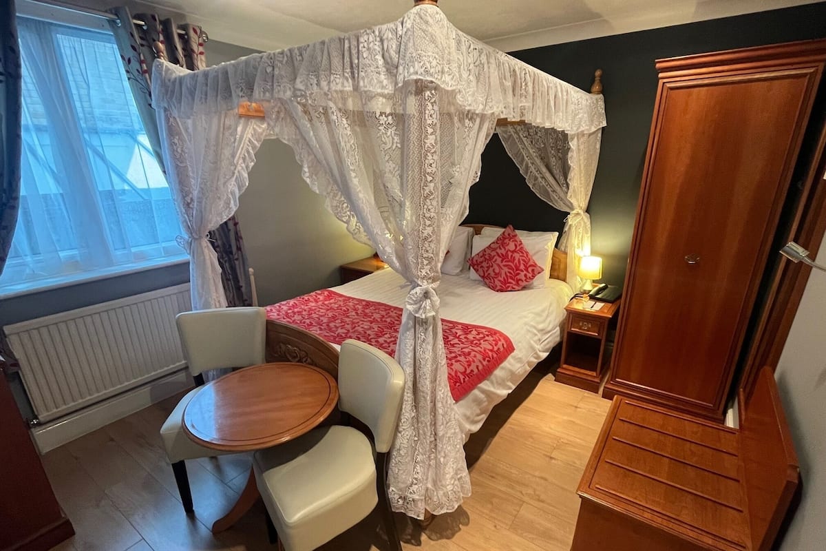 A bedroom with a four-poster bed, a bedside table with a lamp, a wooden wardrobe, a round wooden table with two chairs, and a window with curtains.