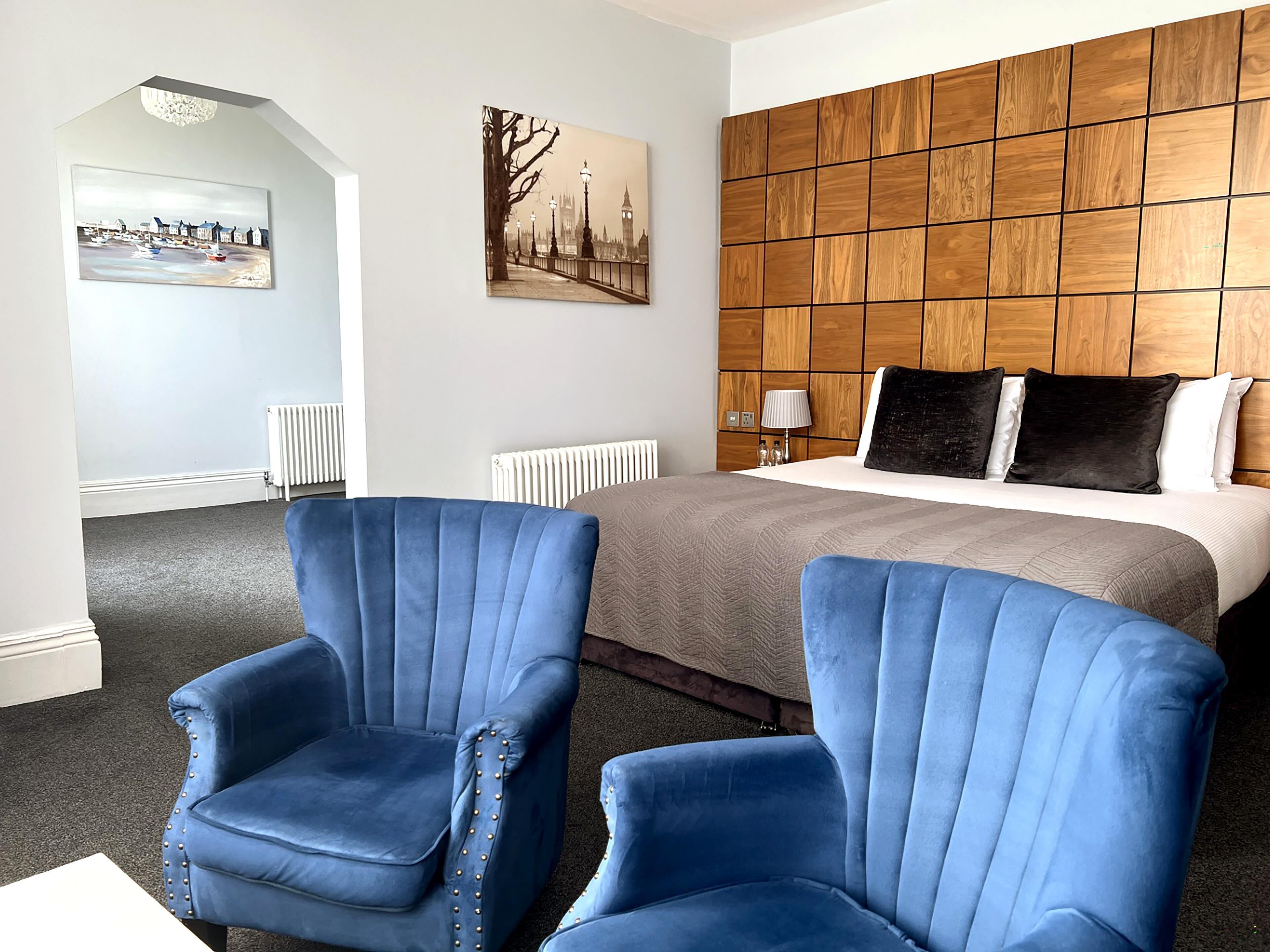 Een hotelkamer met een groot bed, twee blauwe fauteuils, een houten hoofdbord en twee schilderijen aan de muur.