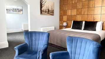 Habitación de hotel con una cama grande, dos sillones azules, un cabecero de madera y dos cuadros en las paredes.