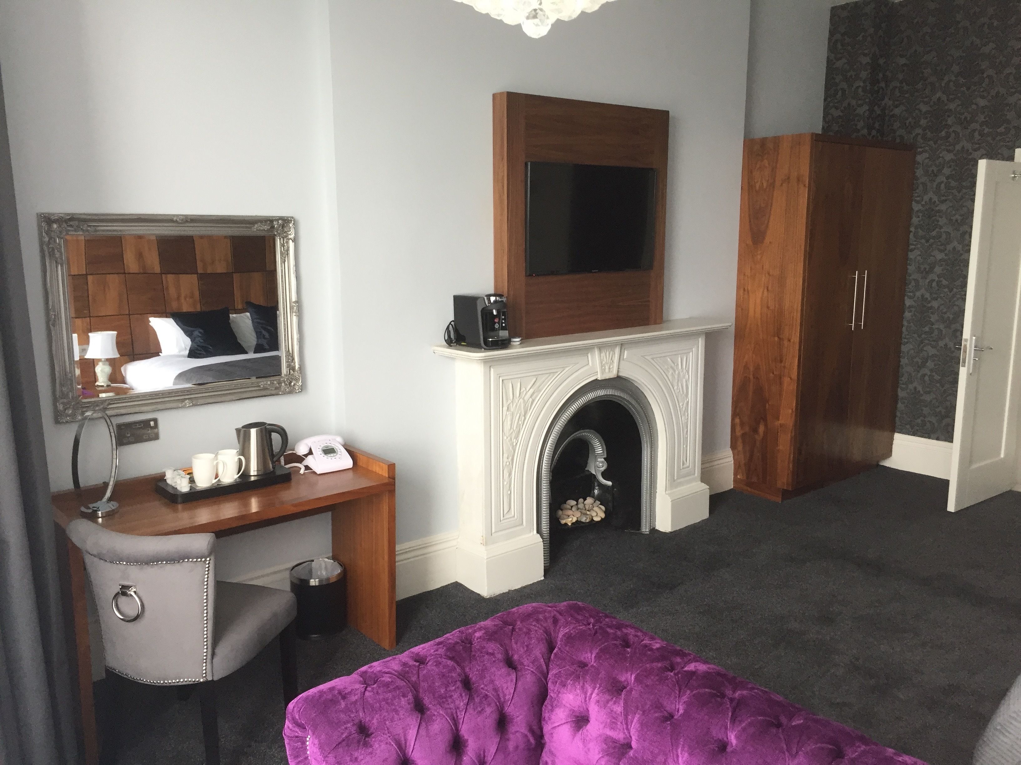 Habitación de hotel con un espejo grande, un escritorio de madera con teléfono y tetera, una chimenea, un sofá morado tapizado y un armario.
