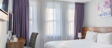 A hotel room with a large bed, a chair, a desk, and a window with curtains.