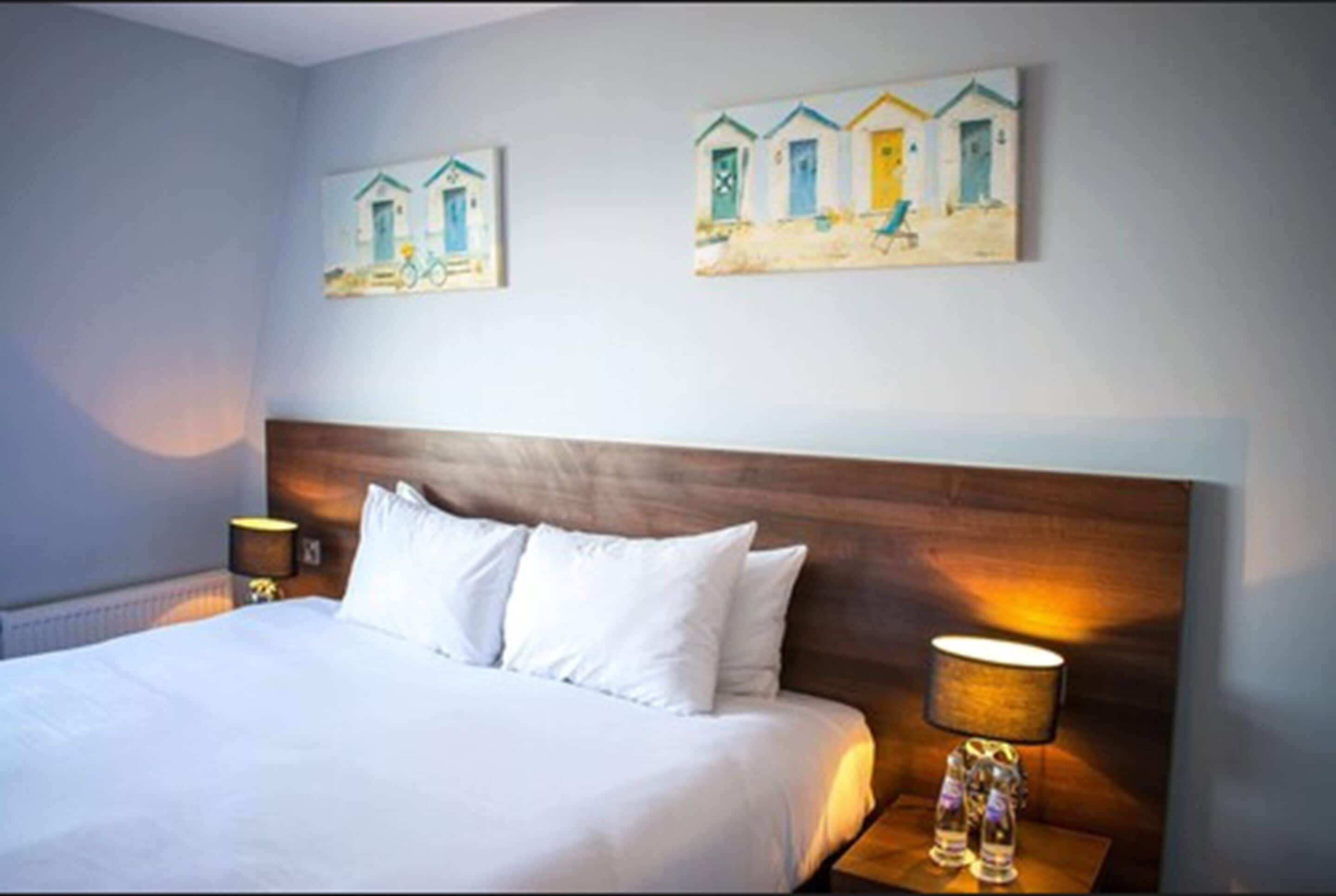 Quarto de hotel com uma cama grande, cabeceira de madeira, duas mesas de cabeceira com abajures e duas pinturas nas paredes retratando cenas de praia.
