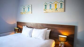 Quarto de hotel com uma cama grande, cabeceira de madeira, duas mesas de cabeceira com abajures e duas pinturas nas paredes retratando cenas de praia.