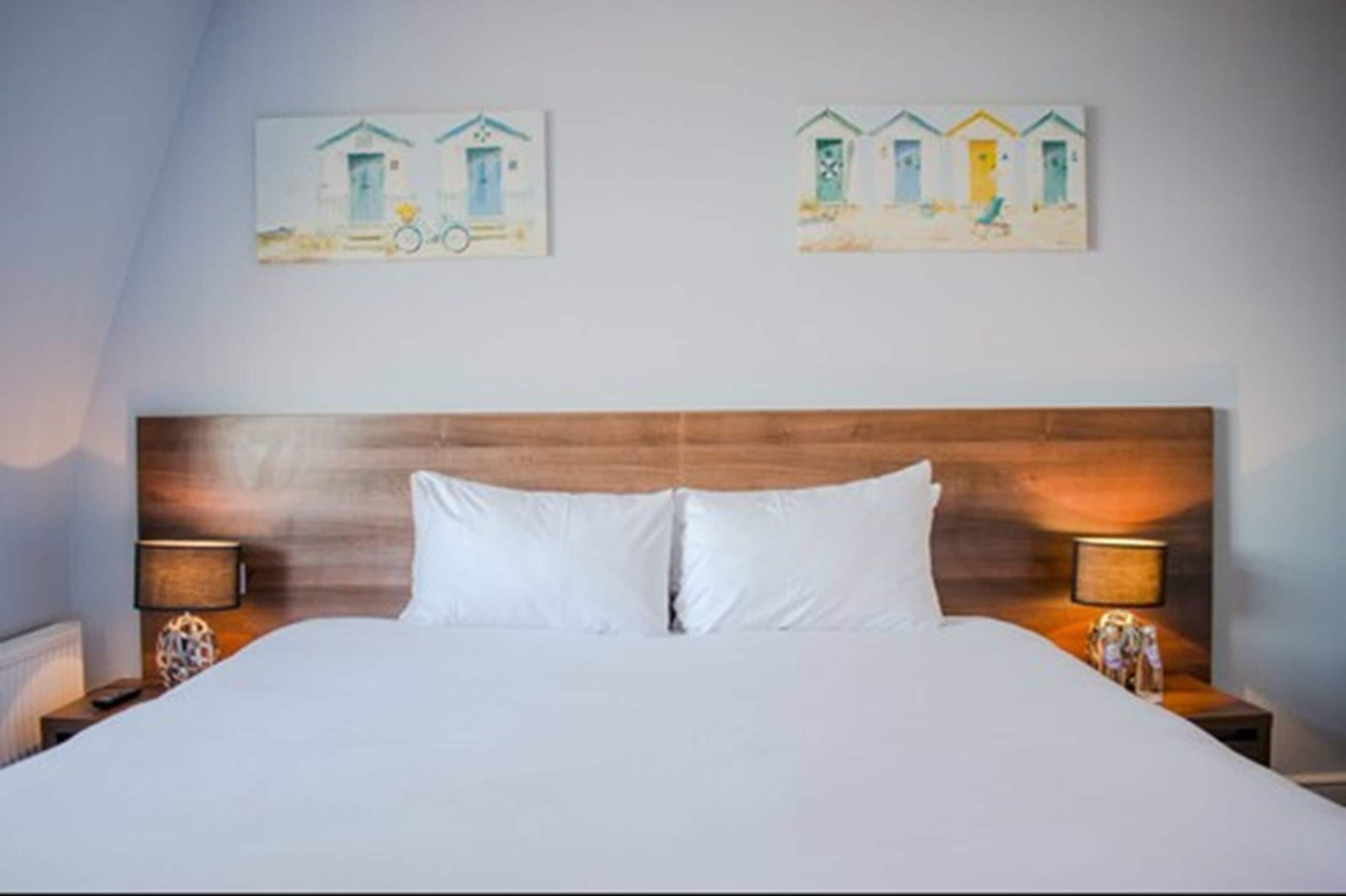 A hotel room with a wooden headboard, white bedding, and two bedside lamps. There are two paintings on the wall depicting beach houses.