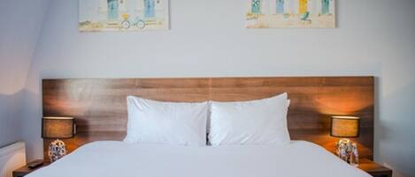 A hotel room with a wooden headboard, white bedding, and two bedside lamps. There are two paintings on the wall depicting beach houses.
