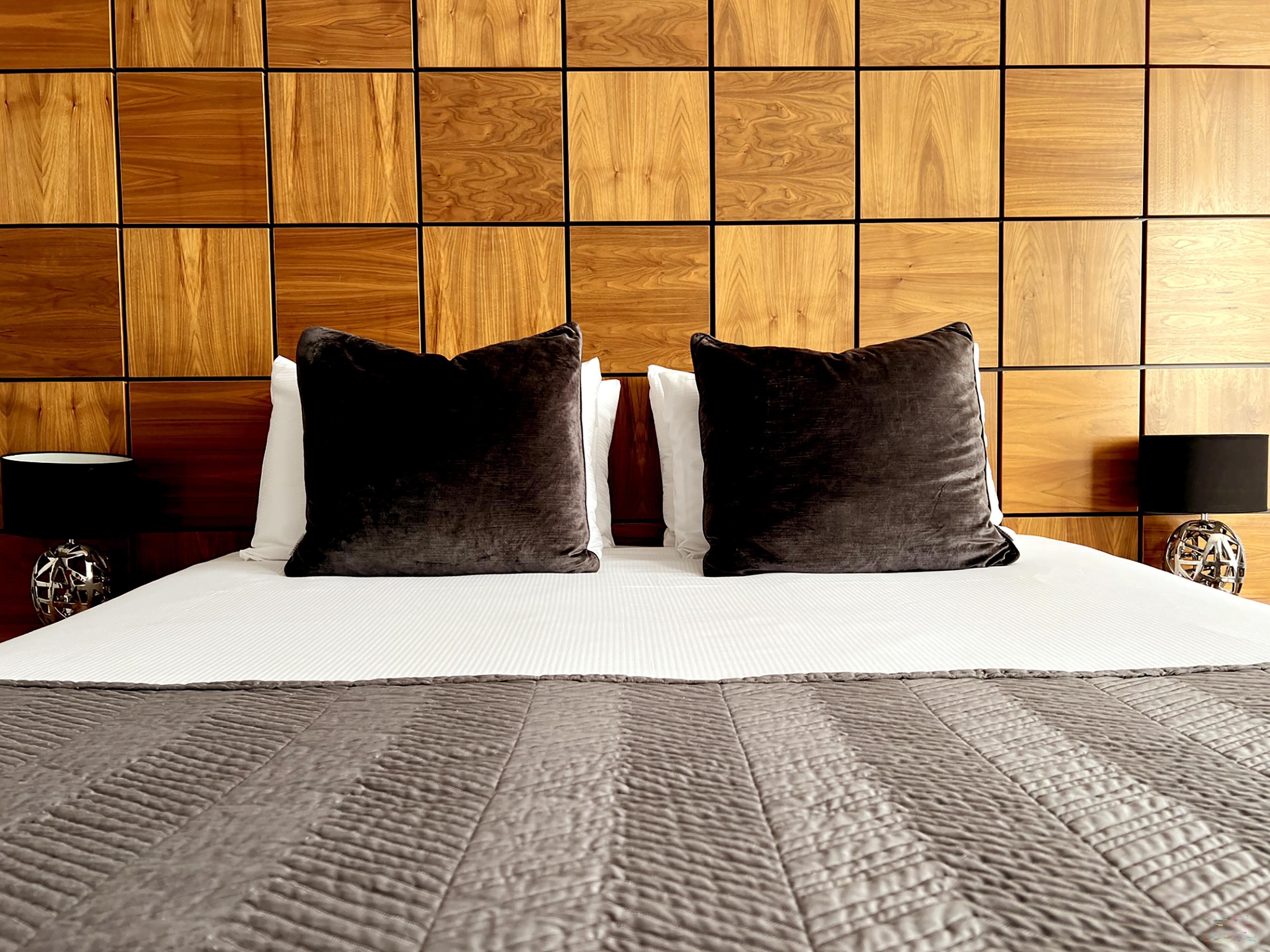 Una cama bien hecha con dos almohadas oscuras, una almohada blanca y una manta gris con textura. Hay dos mesitas de noche con lámparas y objetos decorativos.