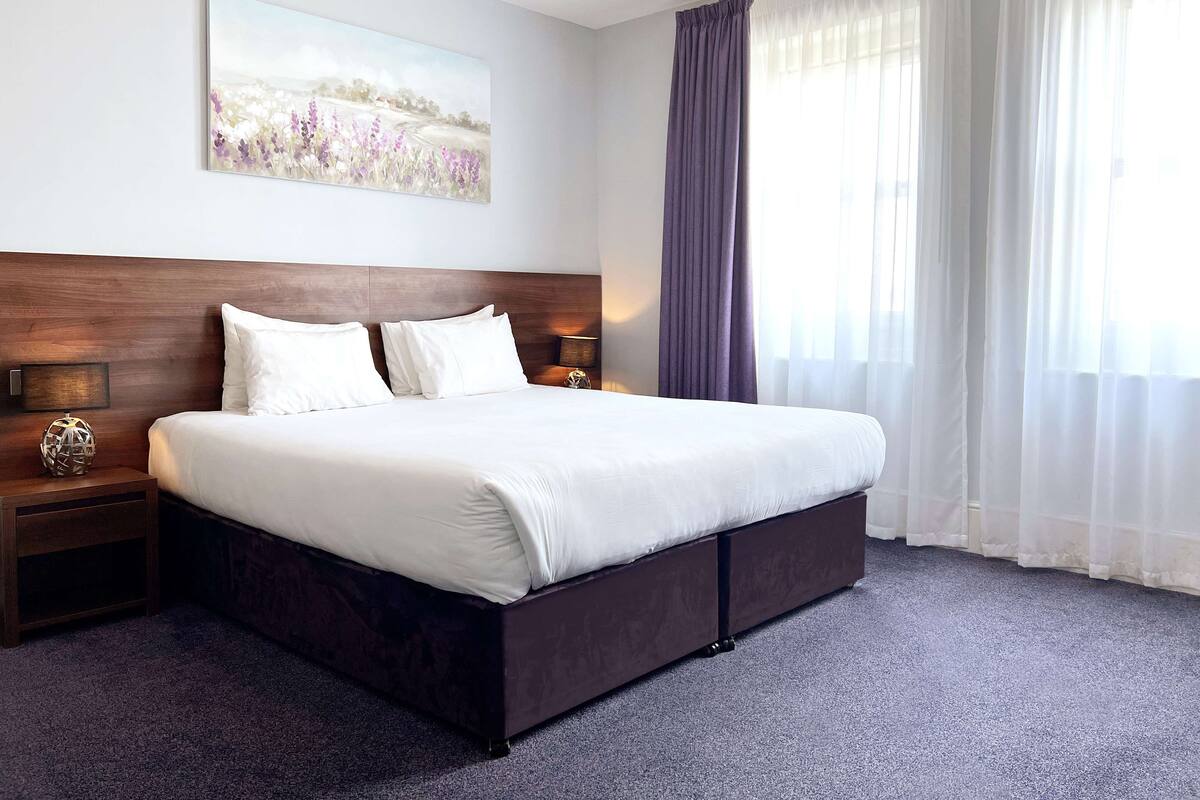 Quarto de hotel com uma cama grande, cabeceira de madeira, duas mesas de cabeceira com luminárias, uma obra de arte na parede e uma janela grande com cortinas leves.