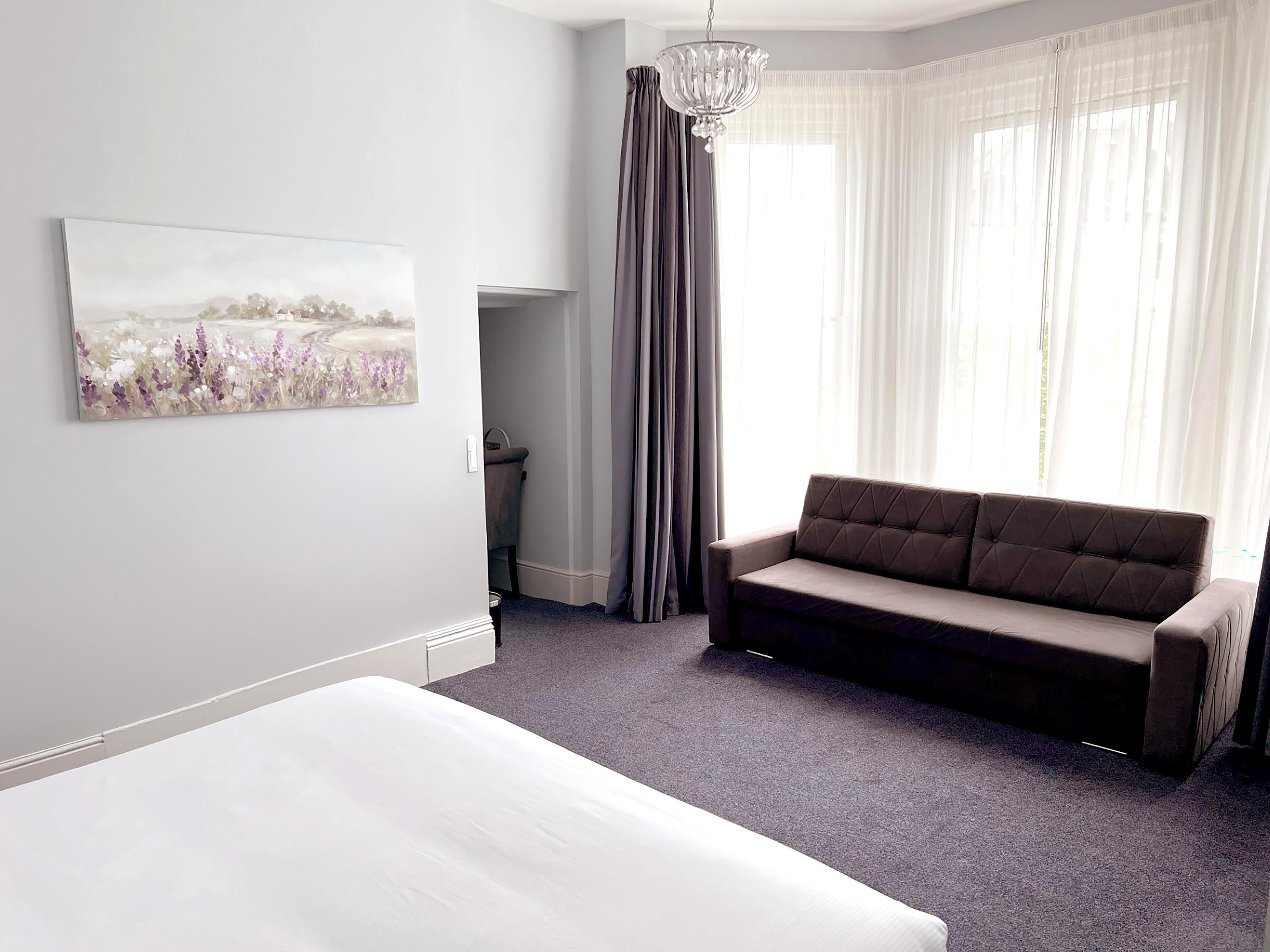 A modern hotel room with a bed, a sofa, a chandelier, and a large window with curtains.