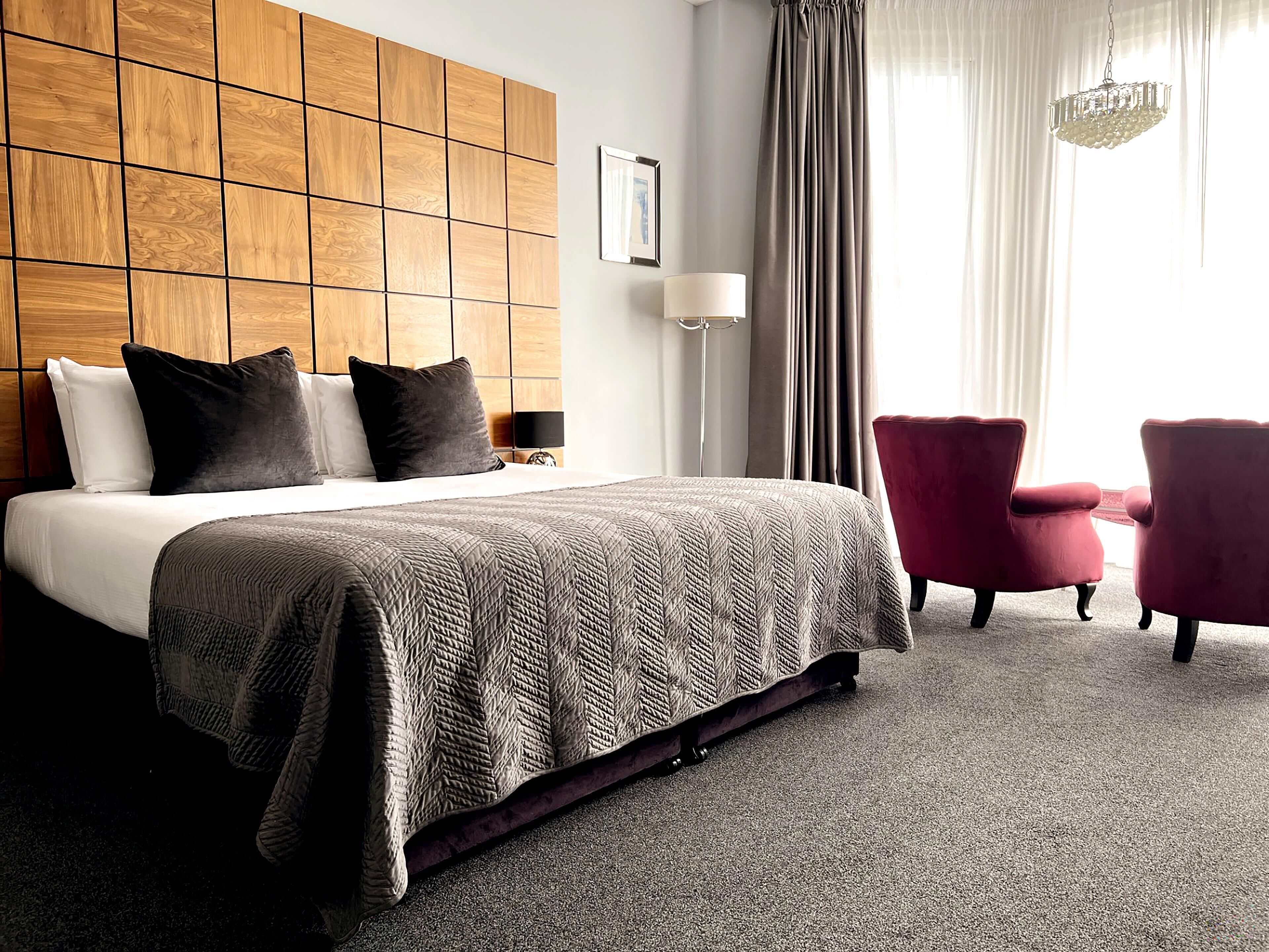 A hotel room with a large bed, a wooden headboard, a red armchair, a grey carpet, and a chandelier.