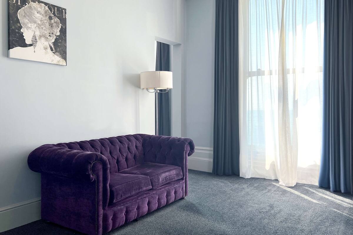 Um quarto com sofá roxo estofado, luminária na parede e uma grande janela com cortinas translúcidas.