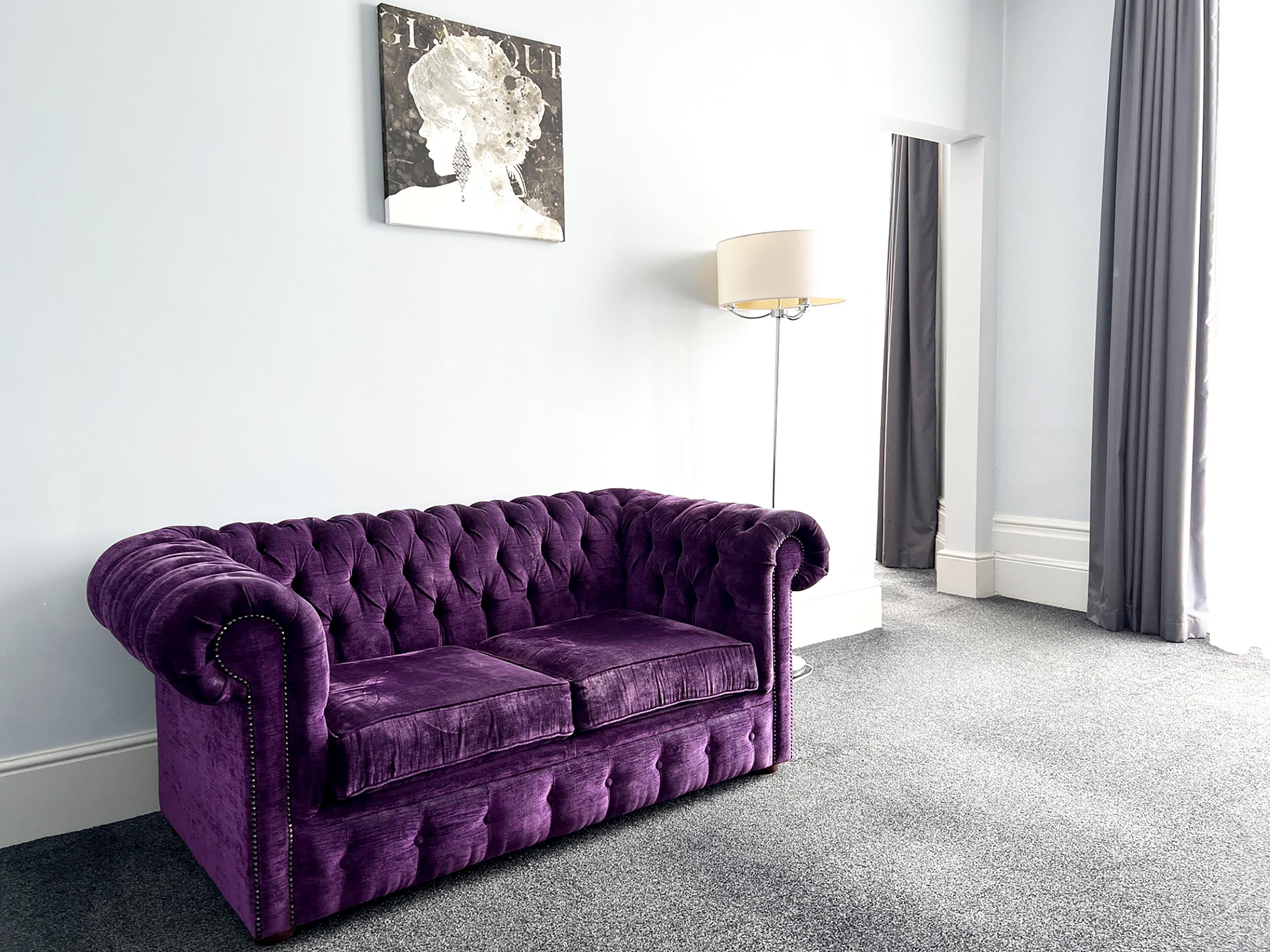 A purple tufted sofa in a hotel room with a white wall, a floor lamp, and a window with curtains.