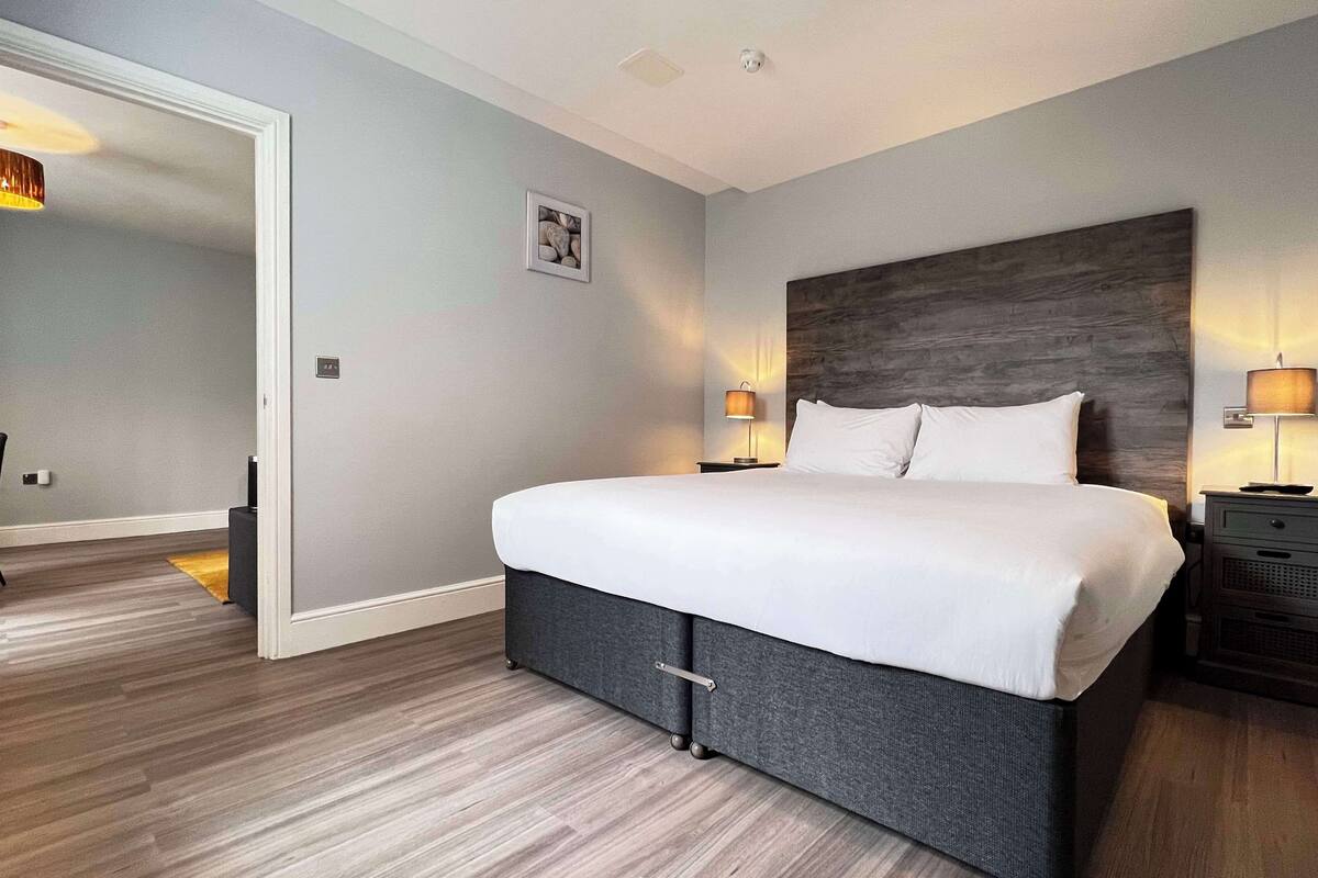 Quarto de hotel moderno com uma cama grande, duas mesas de cabeceira com luminárias, uma cabeceira de madeira e uma pequena imagem na parede.