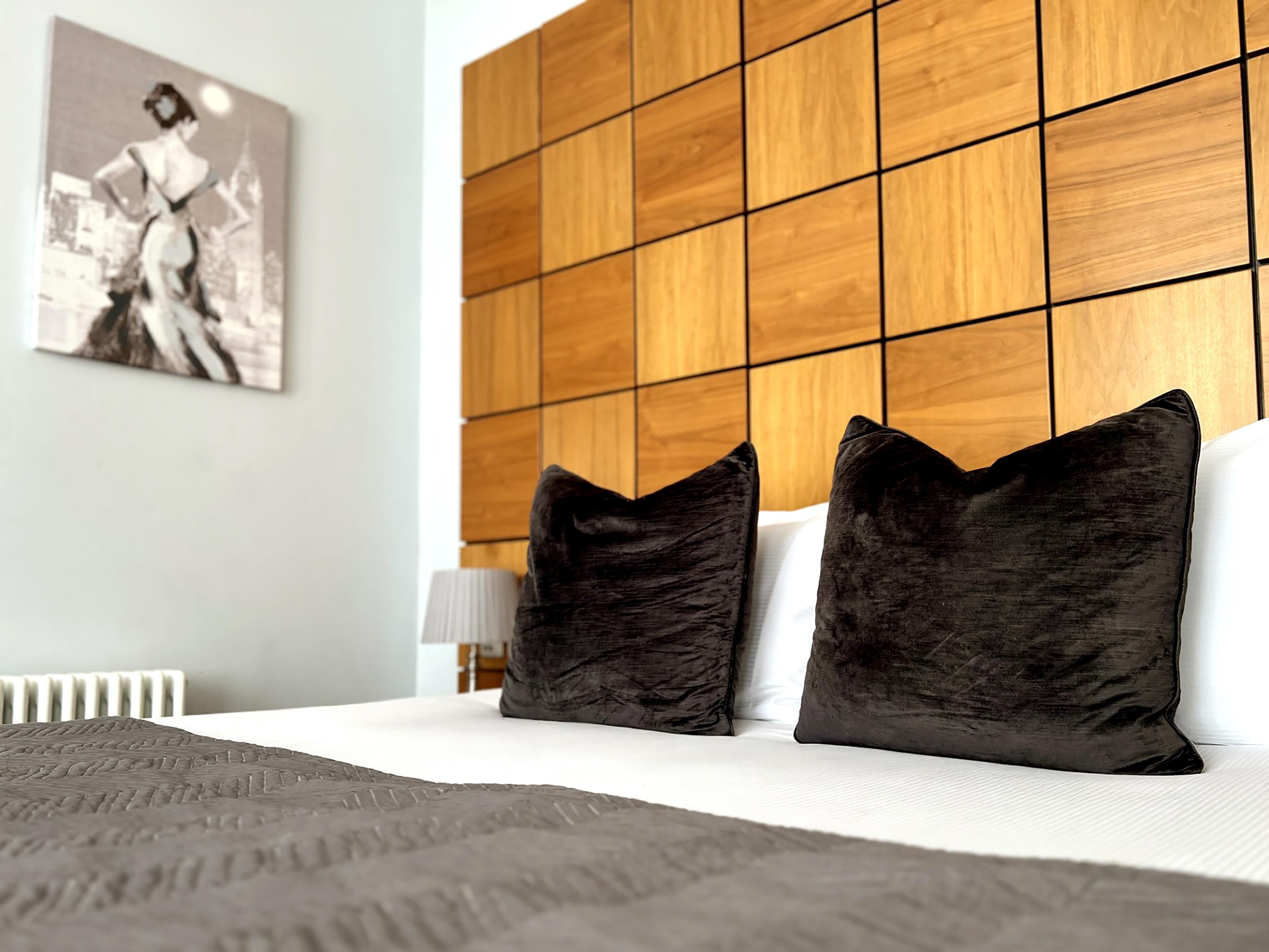 Un lit bien fait, avec des oreillers noirs et une couverture grise, une tête de lit en bois et un tableau encadré accroché au mur.