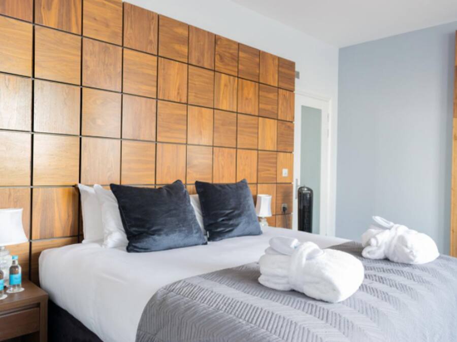A hotel room with a wooden headboard, a neatly made bed with white and dark gray bedding, a bedside table with a lamp and bottles, and a folded towel on the bed.