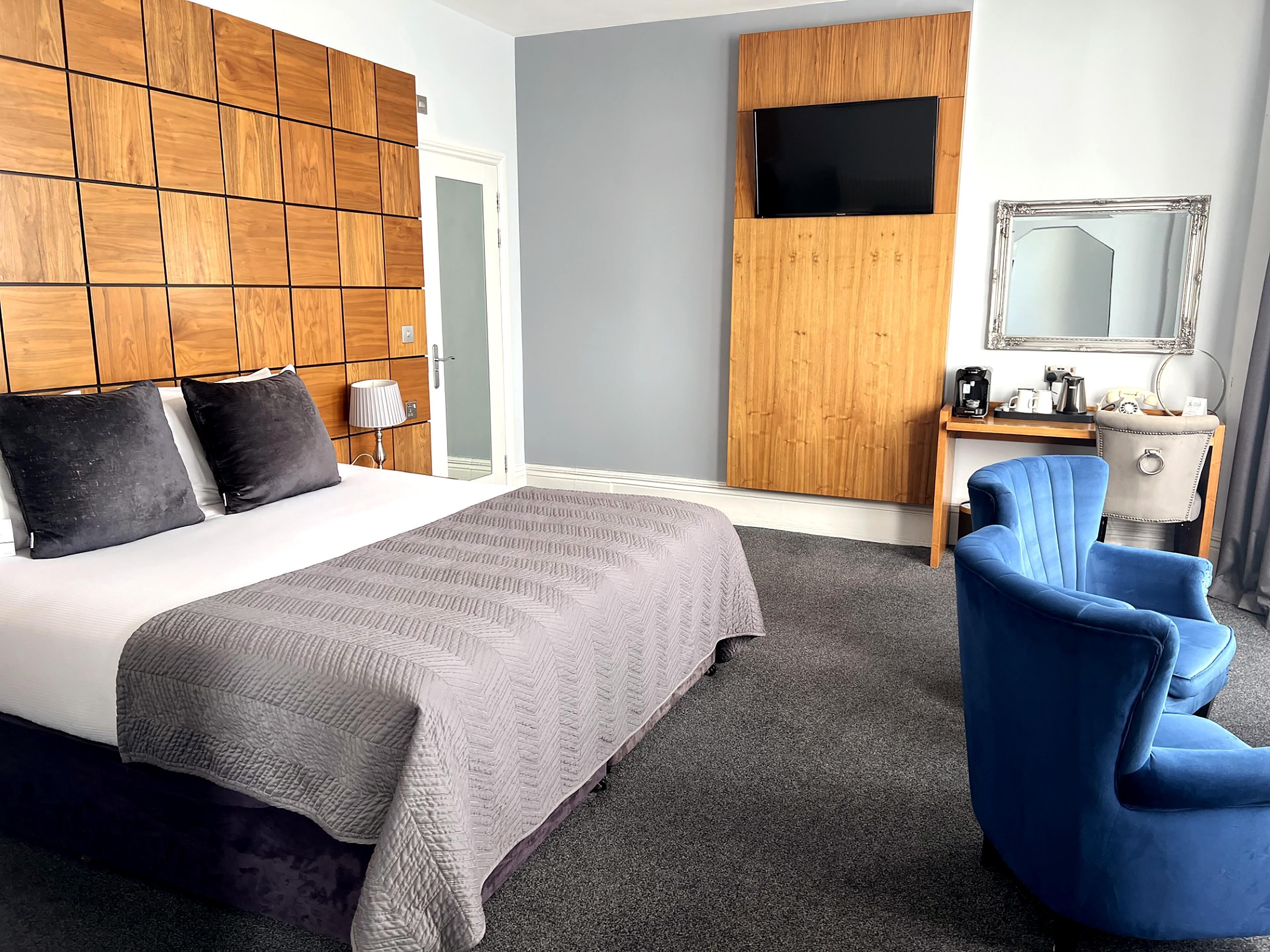 Ein modernes Hotelzimmer mit einem großen Bett, einem hölzernen Kopfteil, einem Schreibtisch mit Spiegel und einem blauen Sessel.
