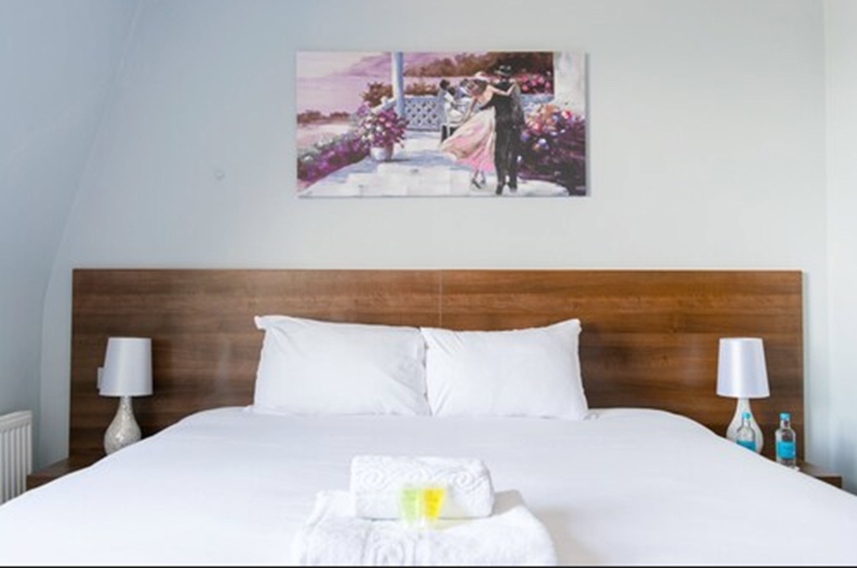 A neatly made bed with white linens, a wooden headboard, and a painting of a couple on the wall.