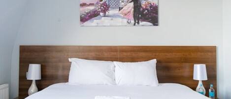 Un lit bien fait, avec du linge de lit blanc, une tête de lit en bois et un tableau représentant un couple accroché au mur.