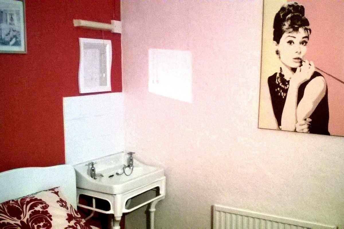 A room with a bed, a small bedside table with a lamp, a wall-mounted picture, and a radiator.