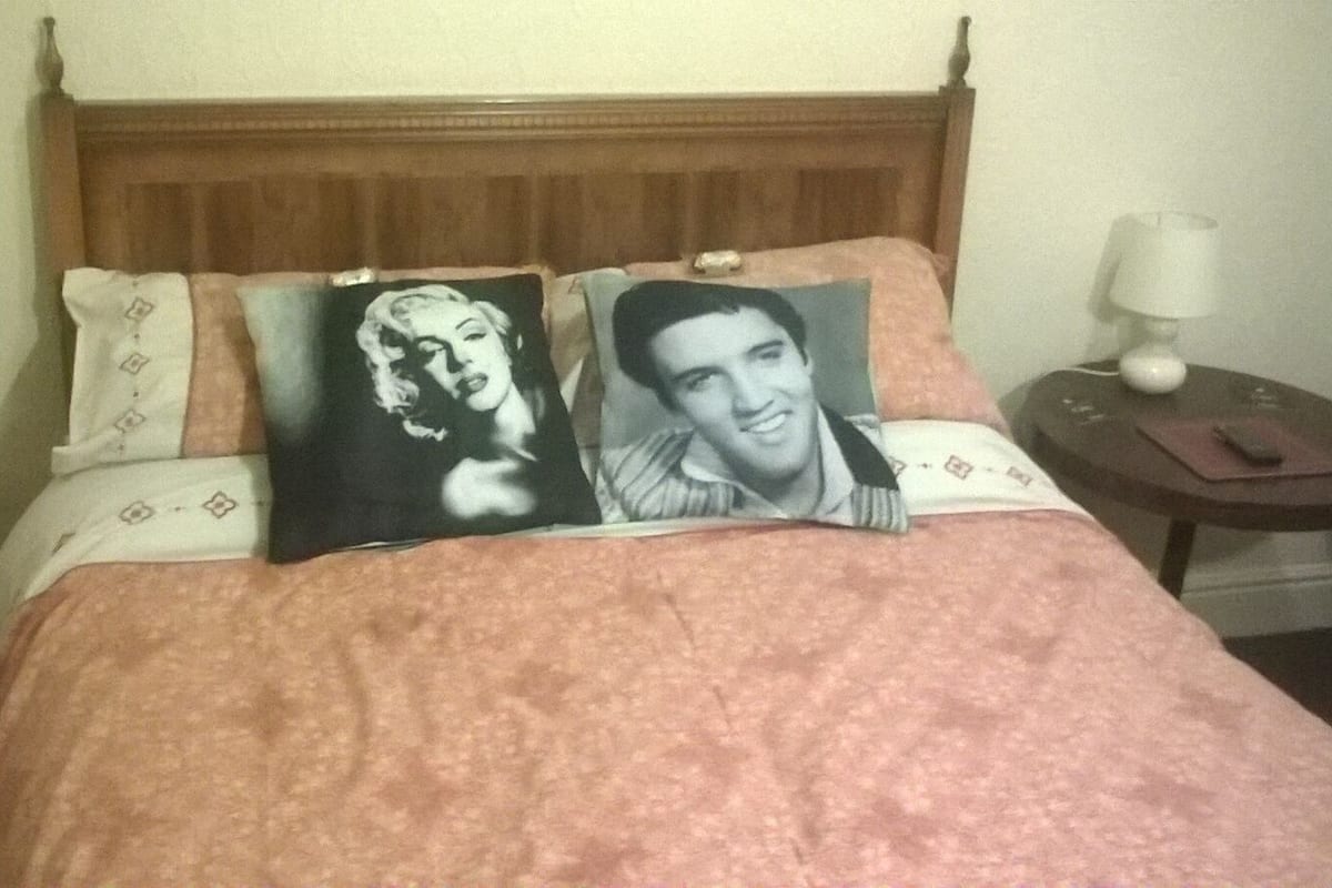 A bed with a pink patterned bedspread, two pillows with printed images, a wooden headboard, a bedside table with a lamp, and a nightstand with a book and a phone.