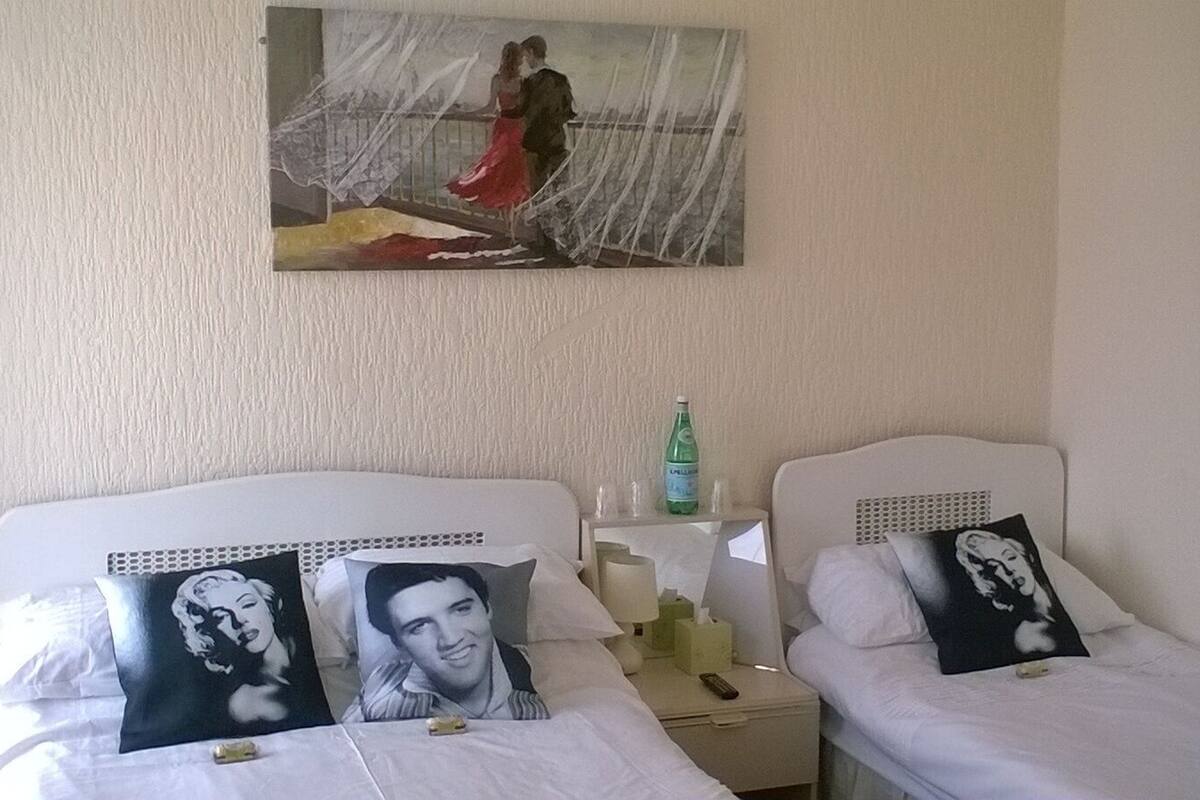 A hotel room with two beds, each with a pillow featuring a black and white portrait. A painting of a couple in a red dress is on the wall above the beds.