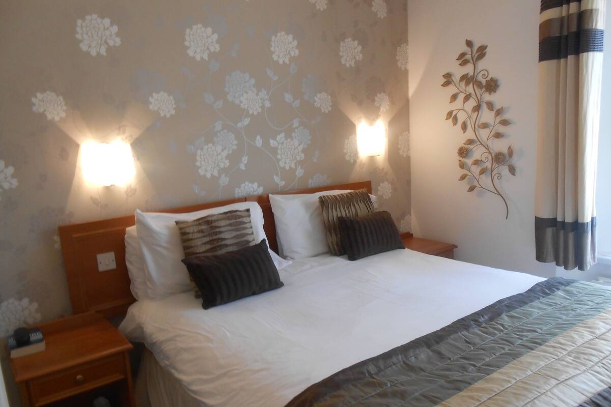 A neatly made bed with white and dark pillows, a wooden headboard, and a wall-mounted light fixture.