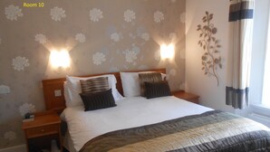 A hotel room with a large bed, two bedside tables, a nightstand, a wall-mounted lamp, and a decorative plant. - Ansteys Lea Guest House (Torquay)