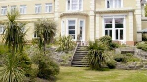 A large, two-story building with a symmetrical facade, multiple windows, and a central entrance with a staircase leading up to it. - Ansteys Lea Guest House (Torquay)