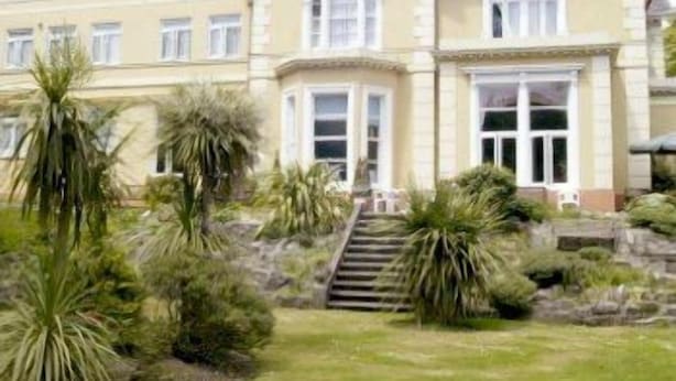 A large, two-story building with a symmetrical facade, multiple windows, and a central entrance with a staircase leading up to it. - Ansteys Lea Guest House (Torquay)