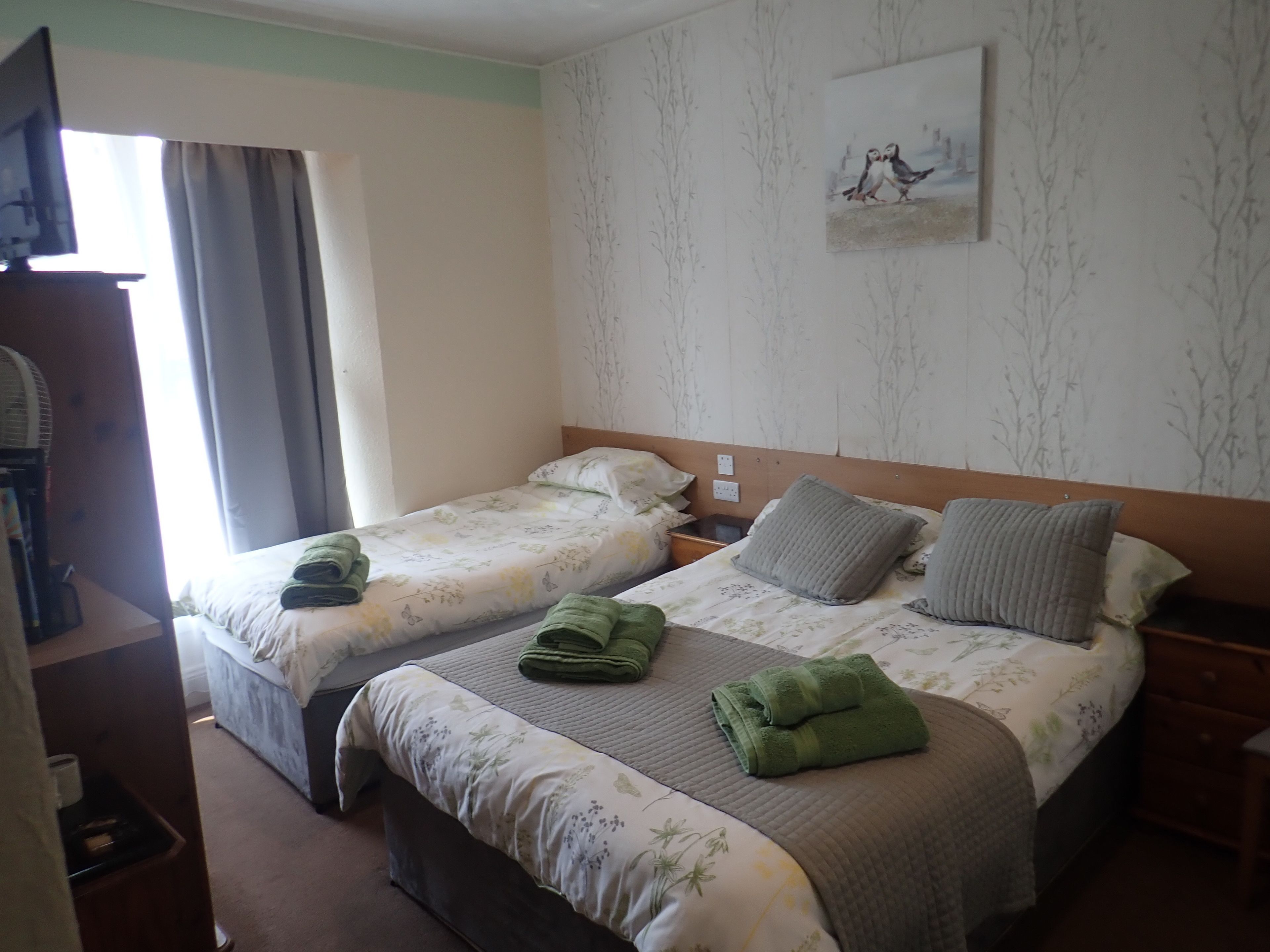Hotellihuone, jossa on kaksi sänkyä. Jokaisessa sängyssä on kukallinen vuodevaate ja koristetyynyt, puinen pääty sekä yöpöytä ja seinälle ripustettu taulu.