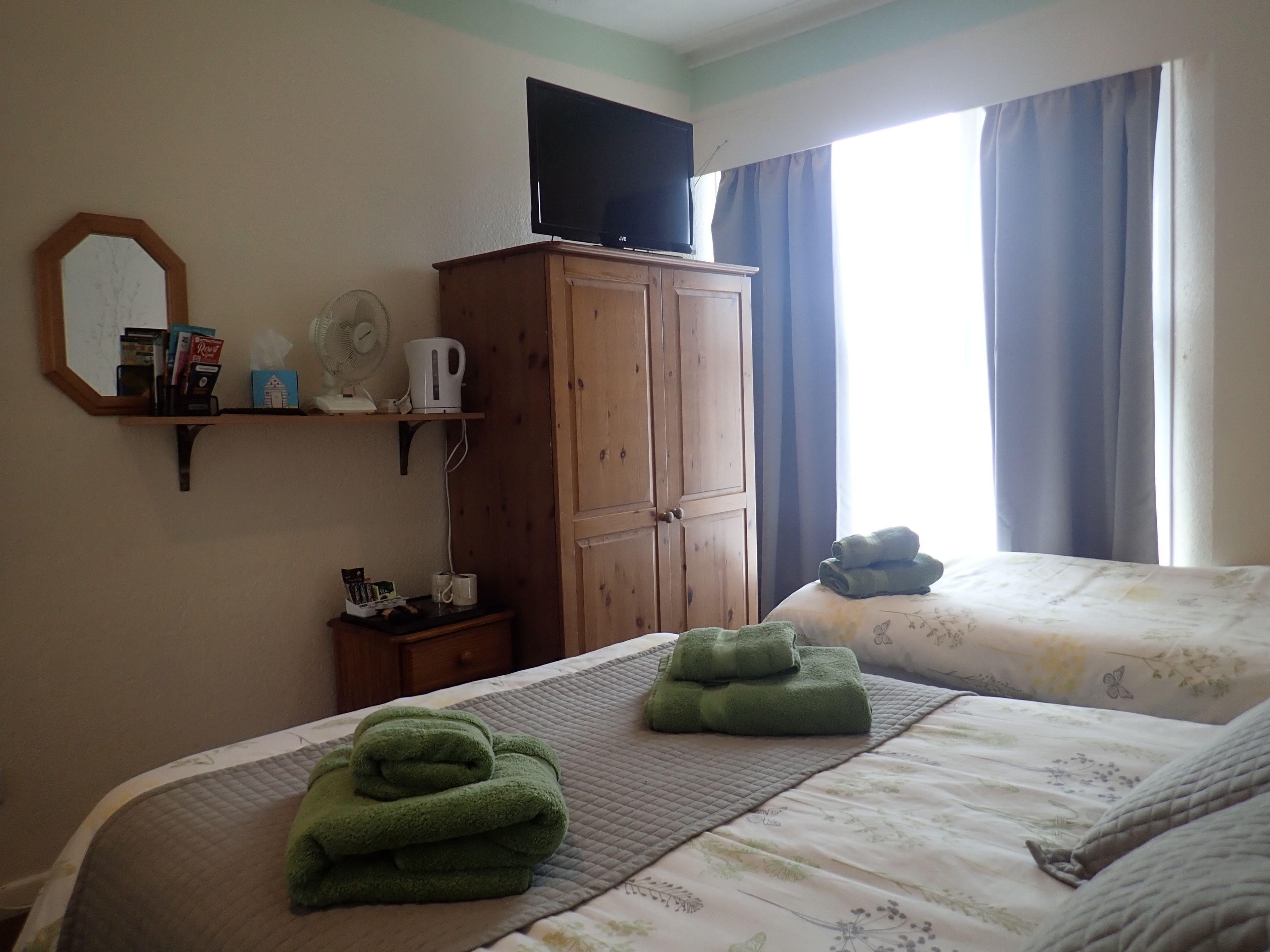 Une chambre avec deux lits, une armoire en bois, une étagère remplie de livres et un ventilateur, ainsi qu’une fenêtre avec des rideaux.